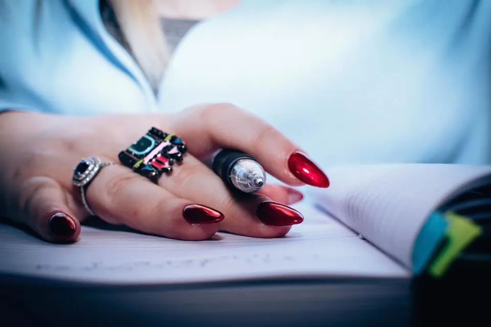 Hand with red manicured nails holding a pen over an open notebook, studying how to become a nail tech in California